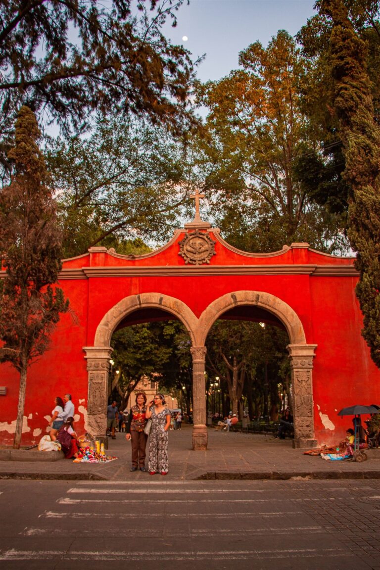 Imagen ilustrativa: acceso peatonal con arco y visitantes en un espacio público; no identifica la sede del evento.
